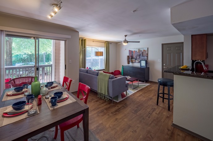 Open-concept apartment at 1250 West furnished with a bed with a dining table with red chairs, a seating area with a couch sliding glass doors to an outdoor balcony, and hardwood floors